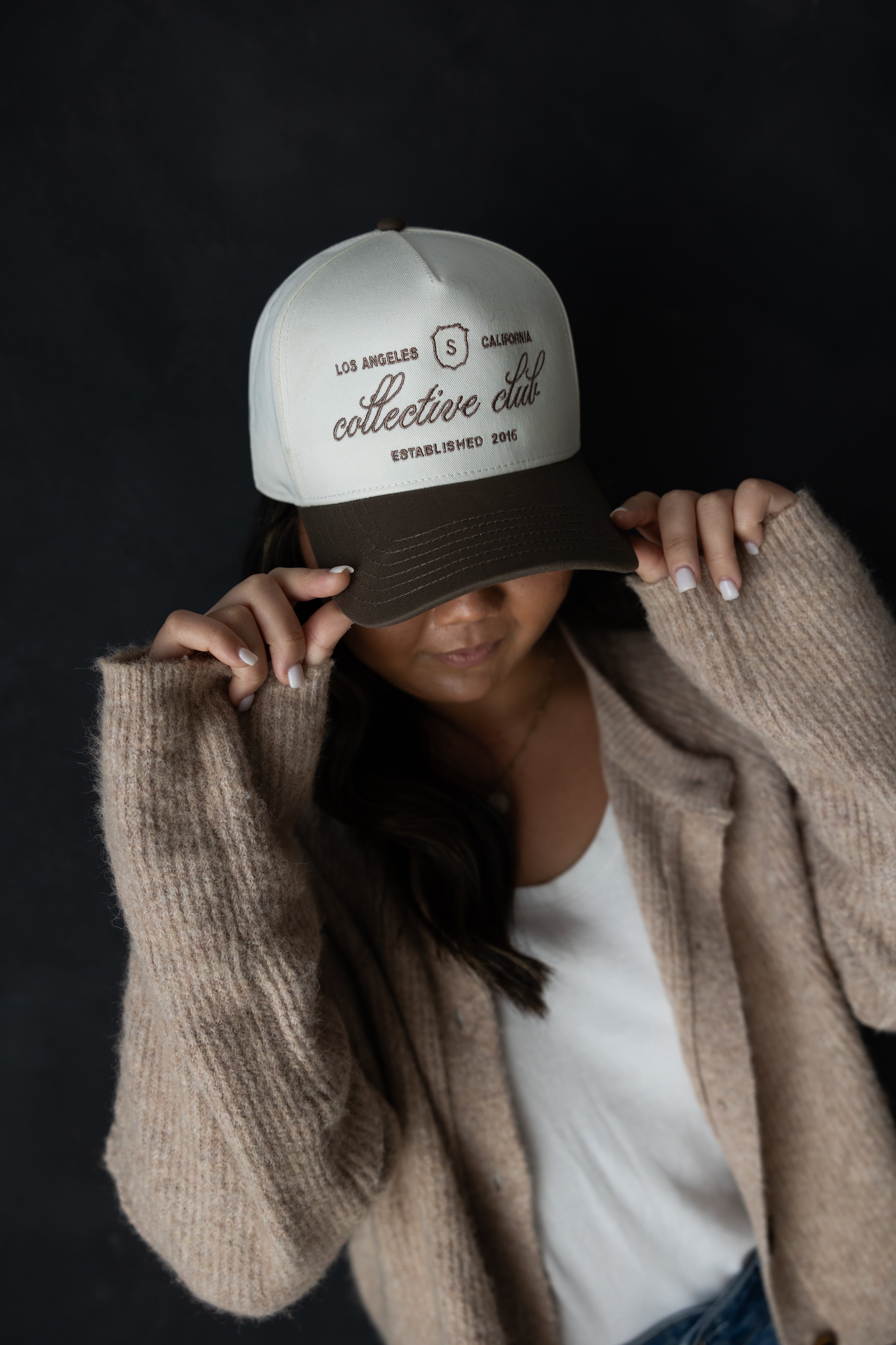 Woman wearing brown and white Collective Club trucker hat by stitched apparel branded as stchd, styled with a soft neutral cardigan and white top, vintage everyday look.