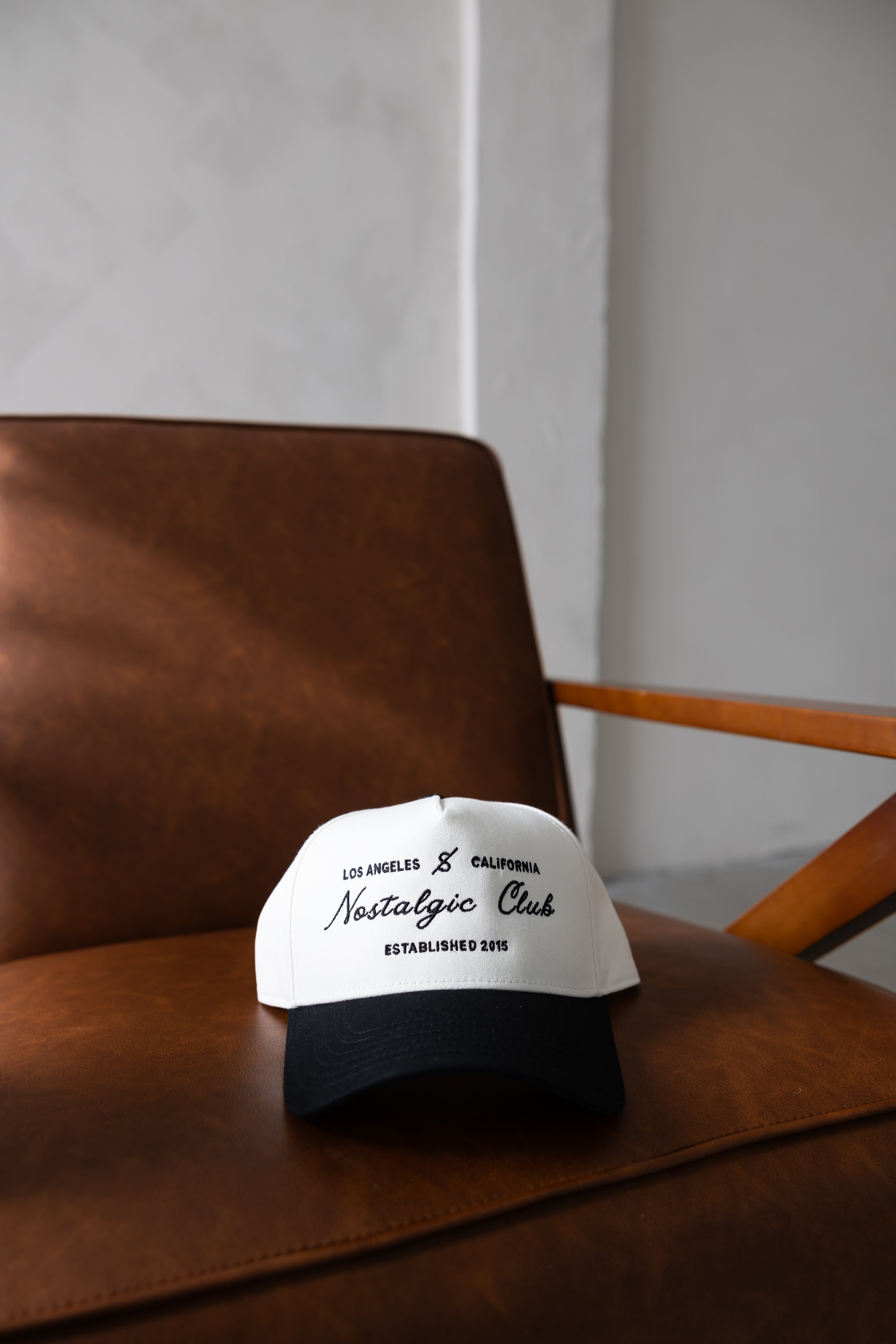 Nostalgic Club black and white vintage trucker hat with Los Angeles embroidery, displayed on a brown leather chair by stitched apparel branded as STCHD