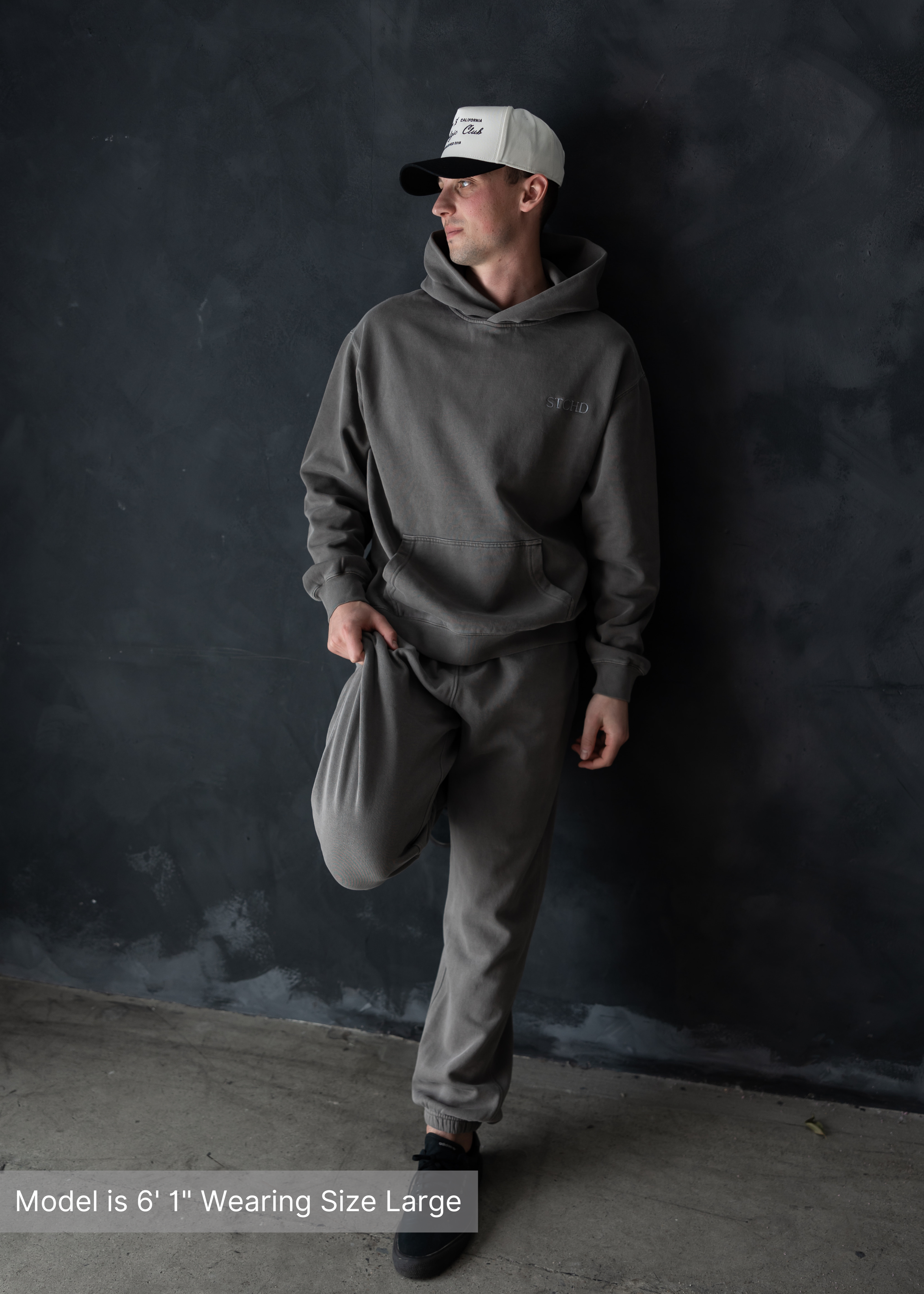 Men’s streetwear look featuring washed grey hoodie and sweatpants set with white and black Los Angeles trucker hat by stitched apparel branded as STCHD, styled against a dark minimalist backdrop.