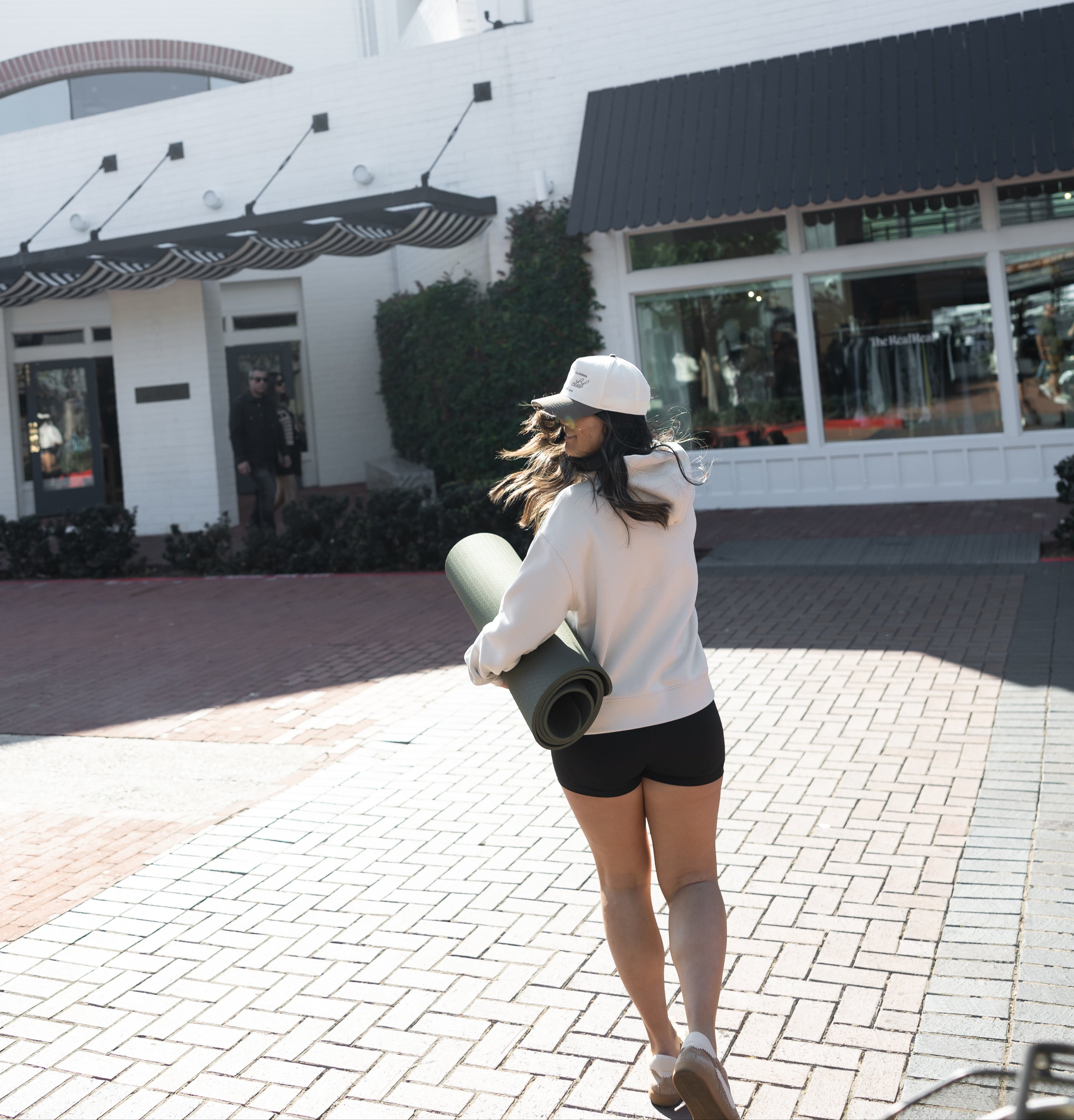 Women’s athleisure outfit with white oversized hoodie, black biker shorts, and Nostalgic Collective hat, walking with yoga mat – active lifestyle and yoga ready look by Stitched Apparel branded as STCHD