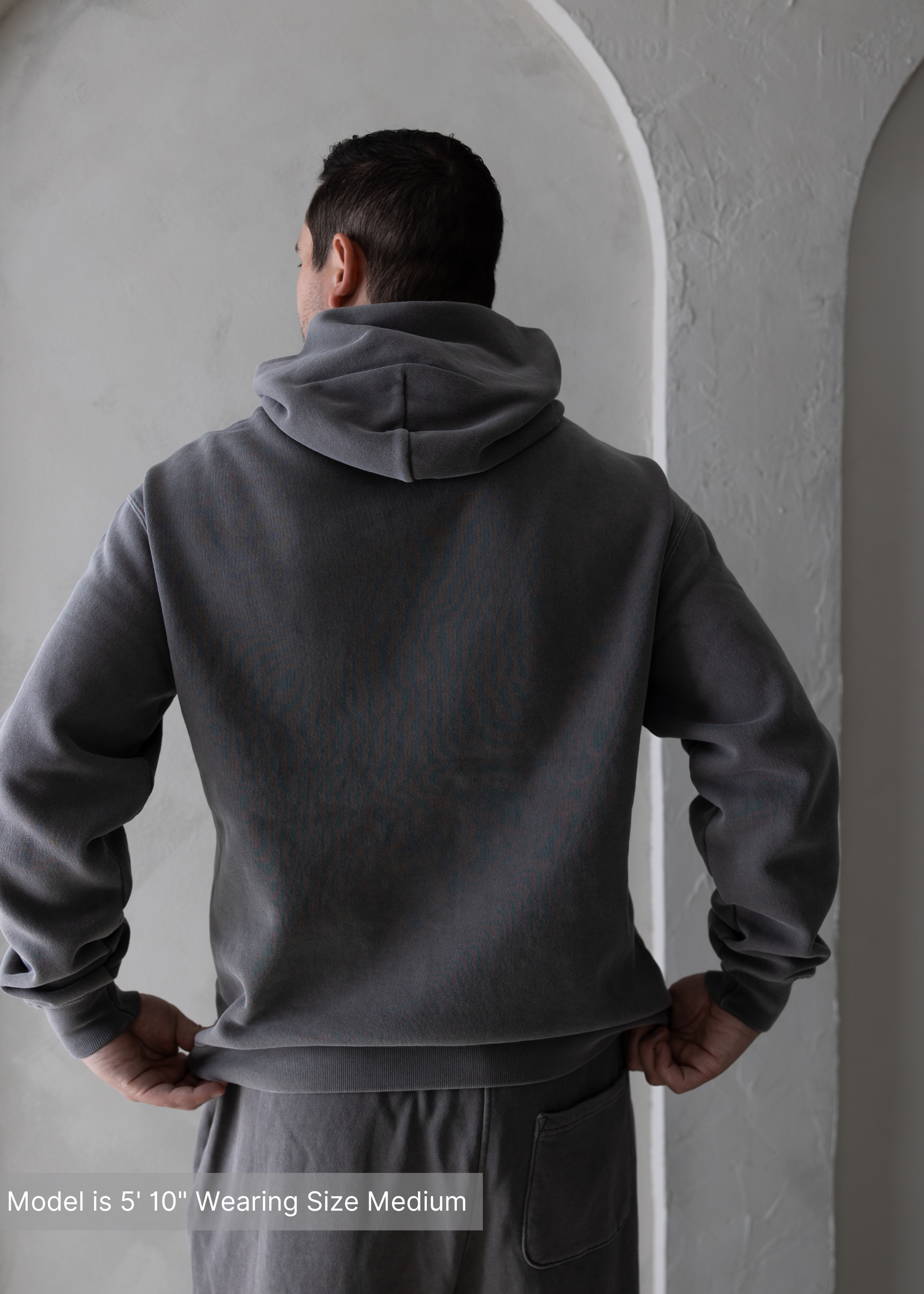 Back view of charcoal men’s hoodie by stitched apparel branded as stchd, hands on hips, standing in front of textured arch wall.
