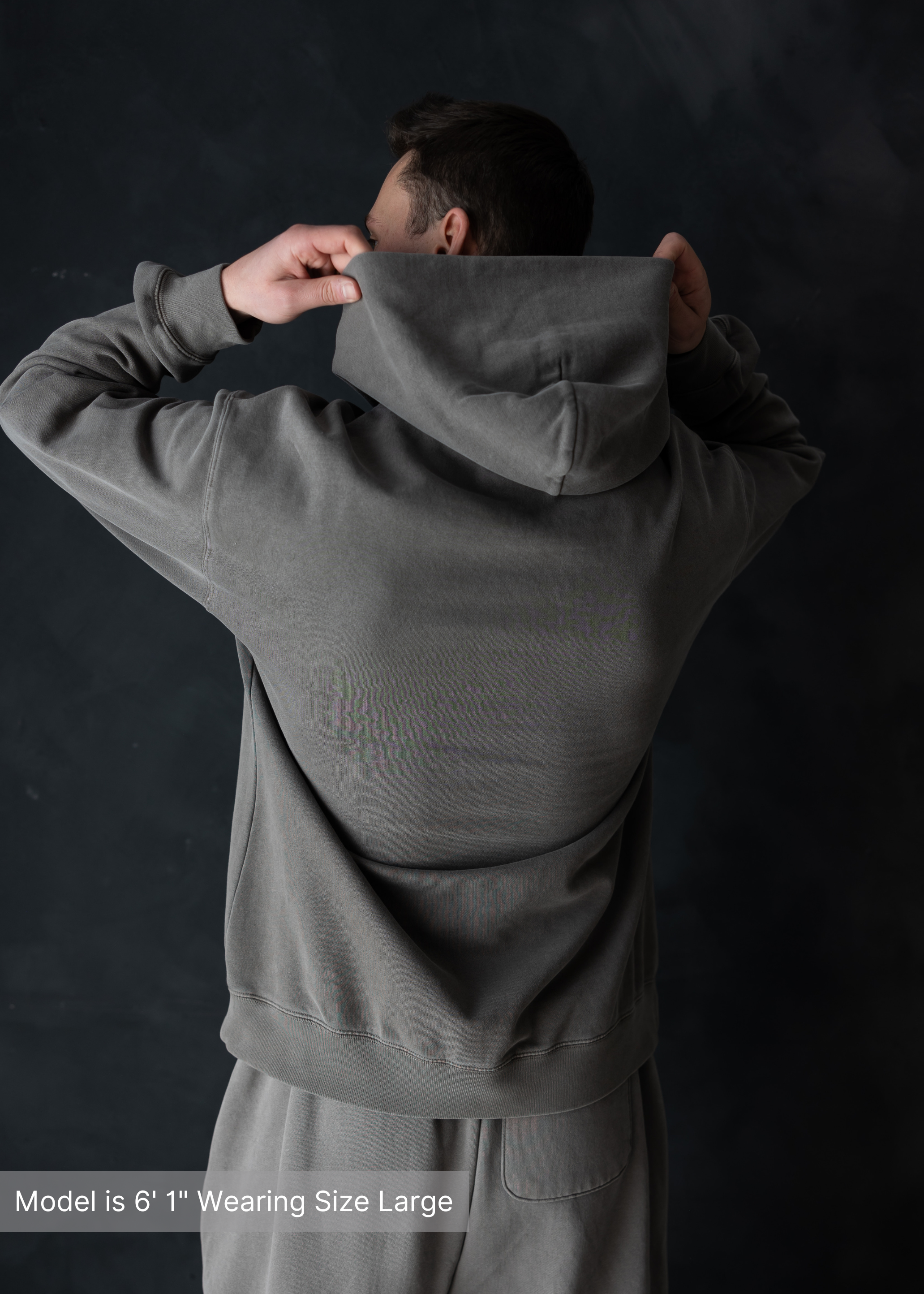 Back view of charcoal hoodie with man lifting hood, dark minimalist background, modern menswear by stitched apparel by stchd
