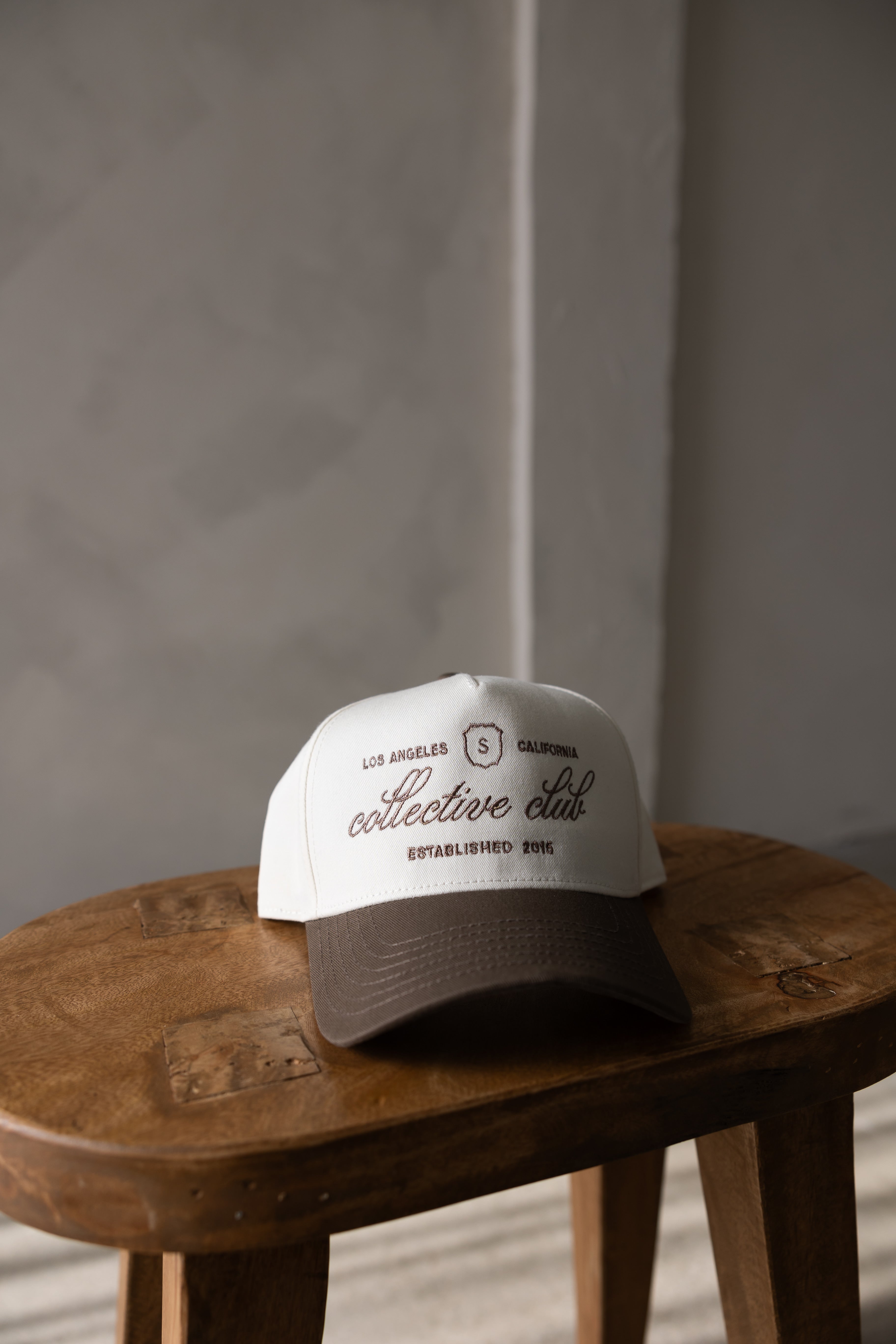 Collective Club brown and white trucker hat with Los Angeles California embroidery, vintage-inspired lifestyle cap on wooden stool by stitched apparel branded as stchd.
