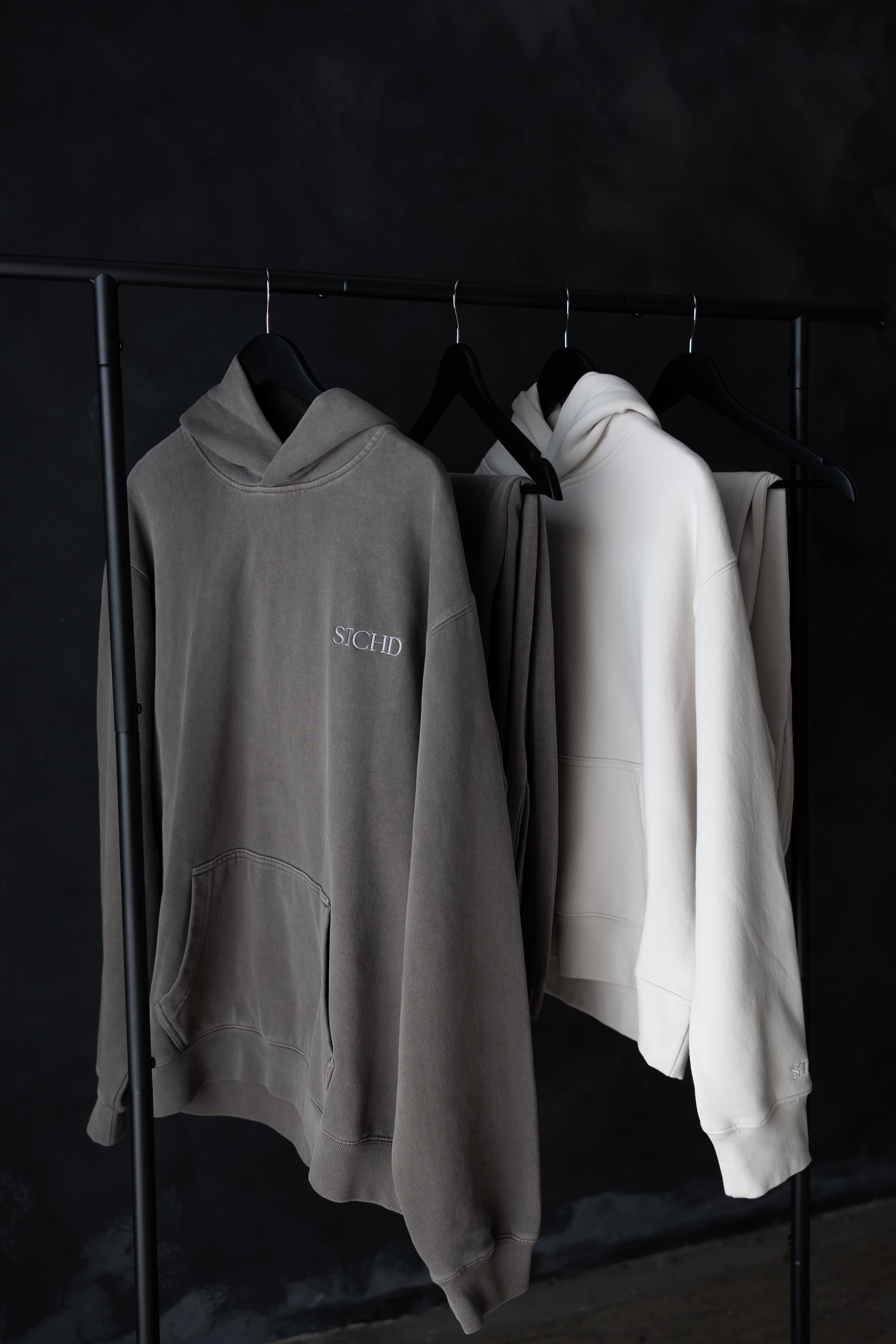 Charcoal and cream STCHD hoodies hanging on black rack, minimal dark background showcasing lifestyle everyday  essentials