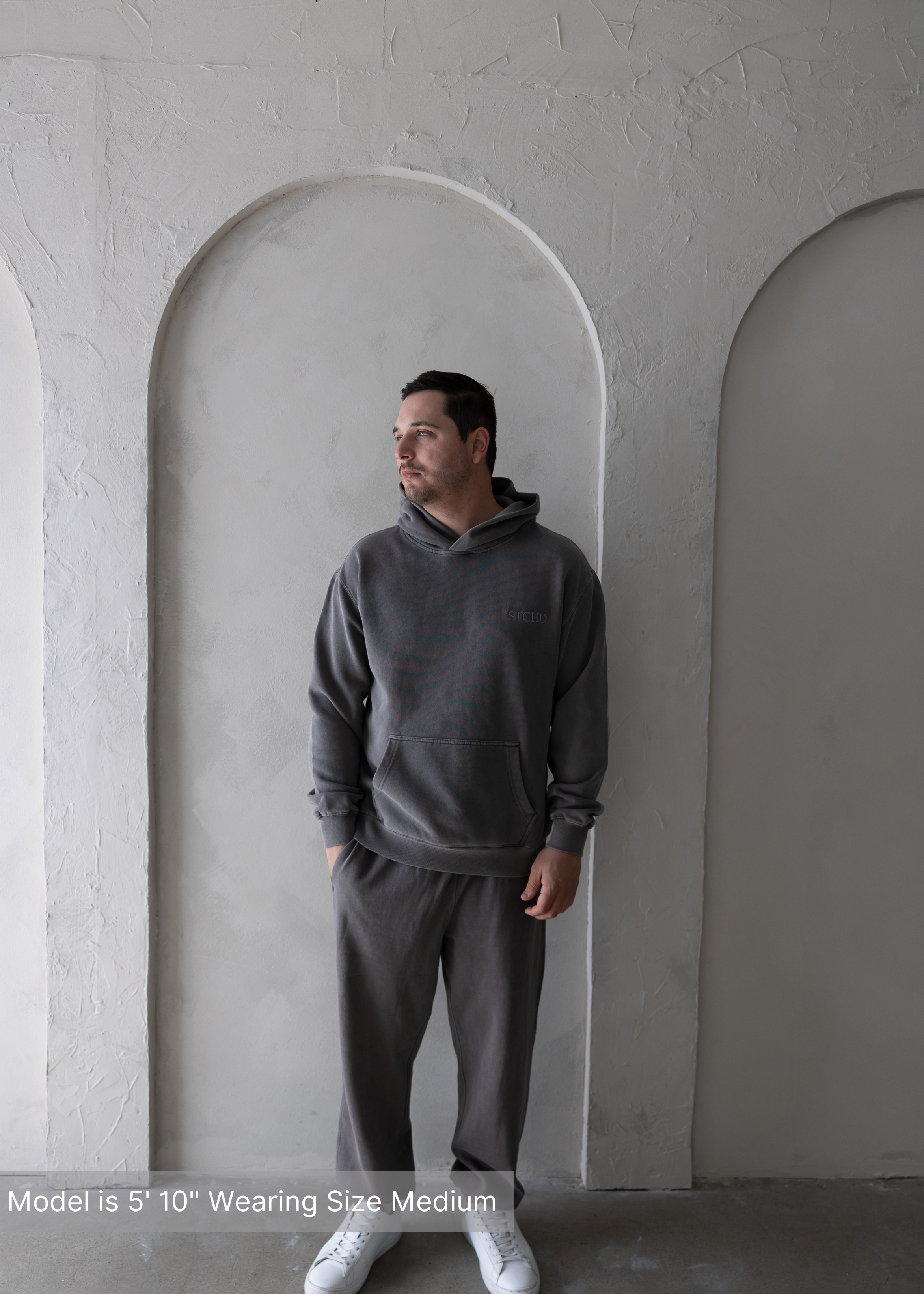 Man standing in washed grey hoodie and jogger sweatpants set by stitched apparel branded as stchd, neutral tone loungewear.