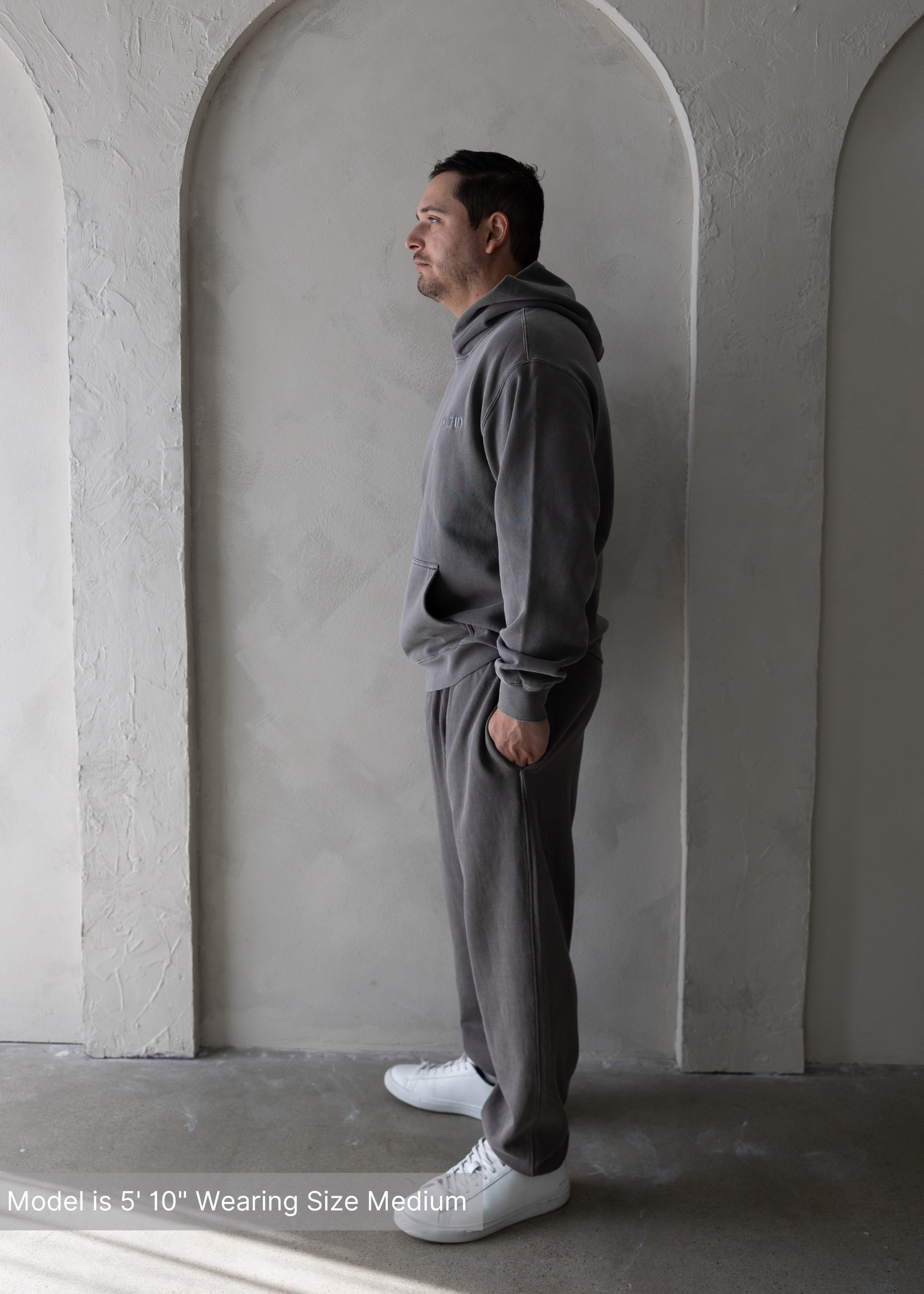 Full body side view of man wearing washed grey hoodie and sweatpants set by stitched apparel branded as stchd against arched neutral wall