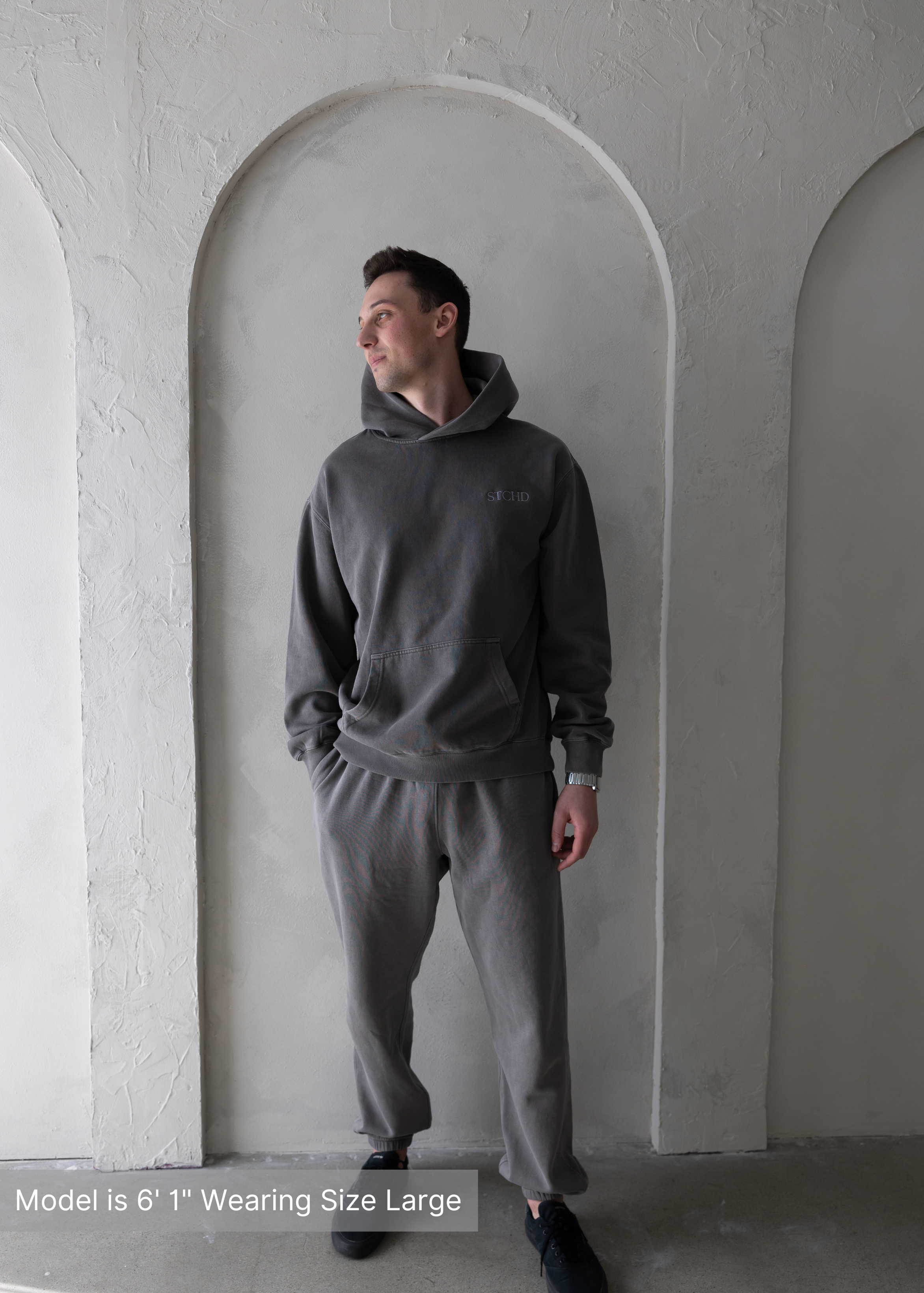 Man wearing washed grey hoodie and sweatpants set by stitched apparel branded as stchd, front view with smile, model is 6’1” and wearing size large