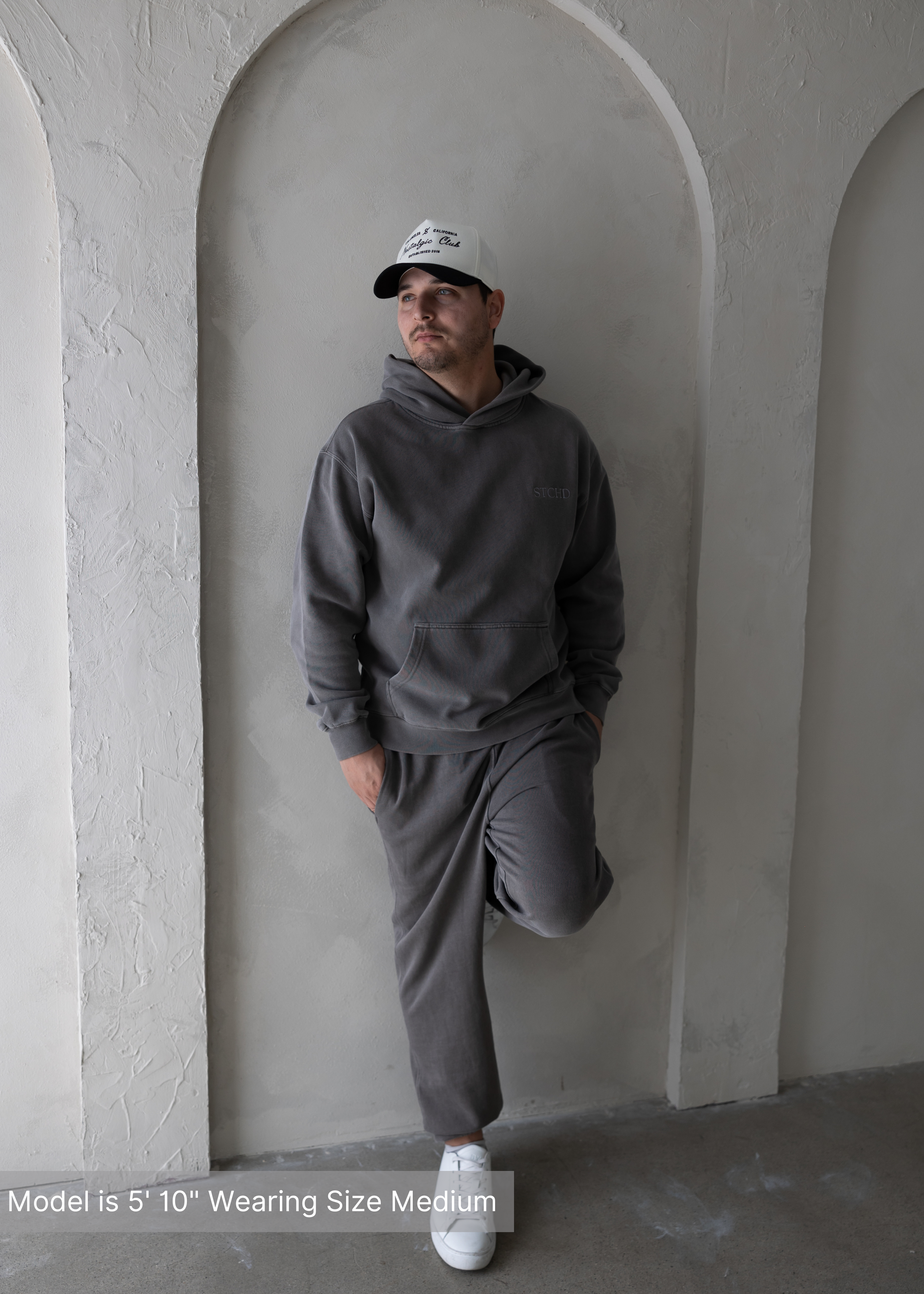 Man wearing a washed grey hoodie and sweatpants set by stitched apparel branded as STCHD paired with a white and black Los Angeles Nostalgic Club trucker hat, leaning against a textured arch wall.
