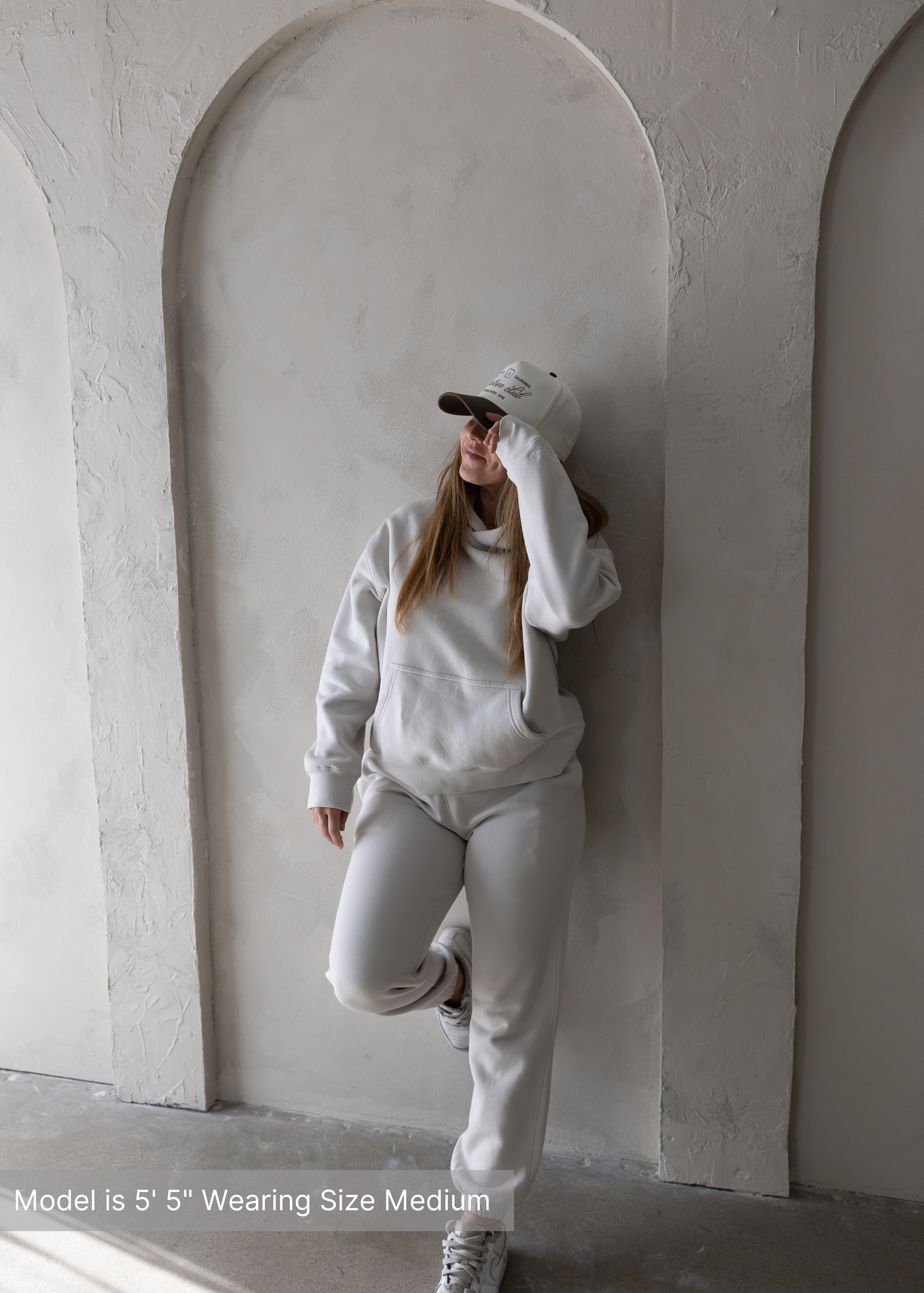 Woman wearing a cozy neutral loungewear set with hoodie and sweatpants, paired with a beige streetwear baseball cap by stitched apparel branded as STCHD, standing against a textured arch wall