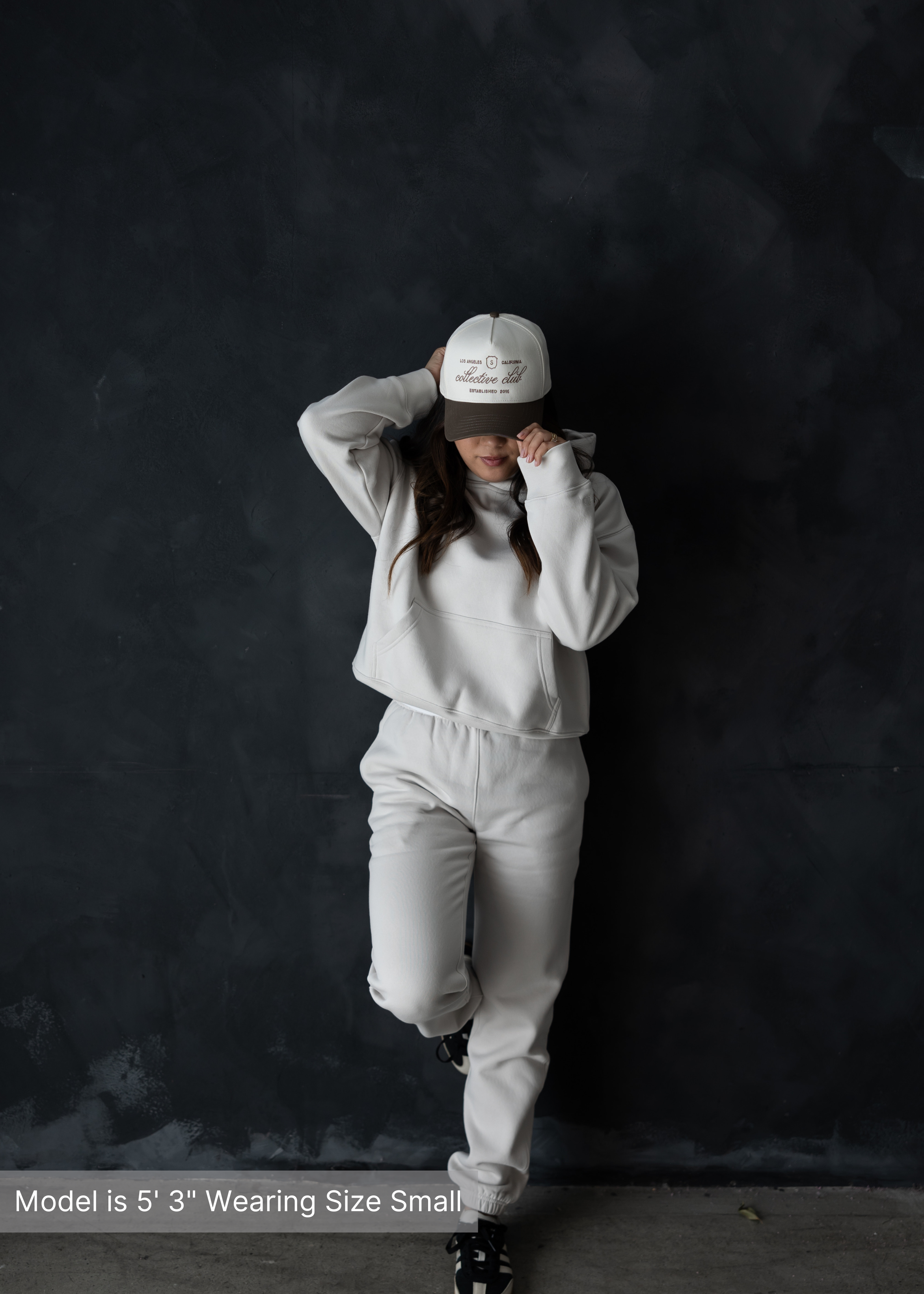Woman wearing a comfortable neutral loungewear set with hoodie and sweatpants, paired with a beige streetwear baseball cap by stitched apparel branded as STCHD, standing against a textured arch wall