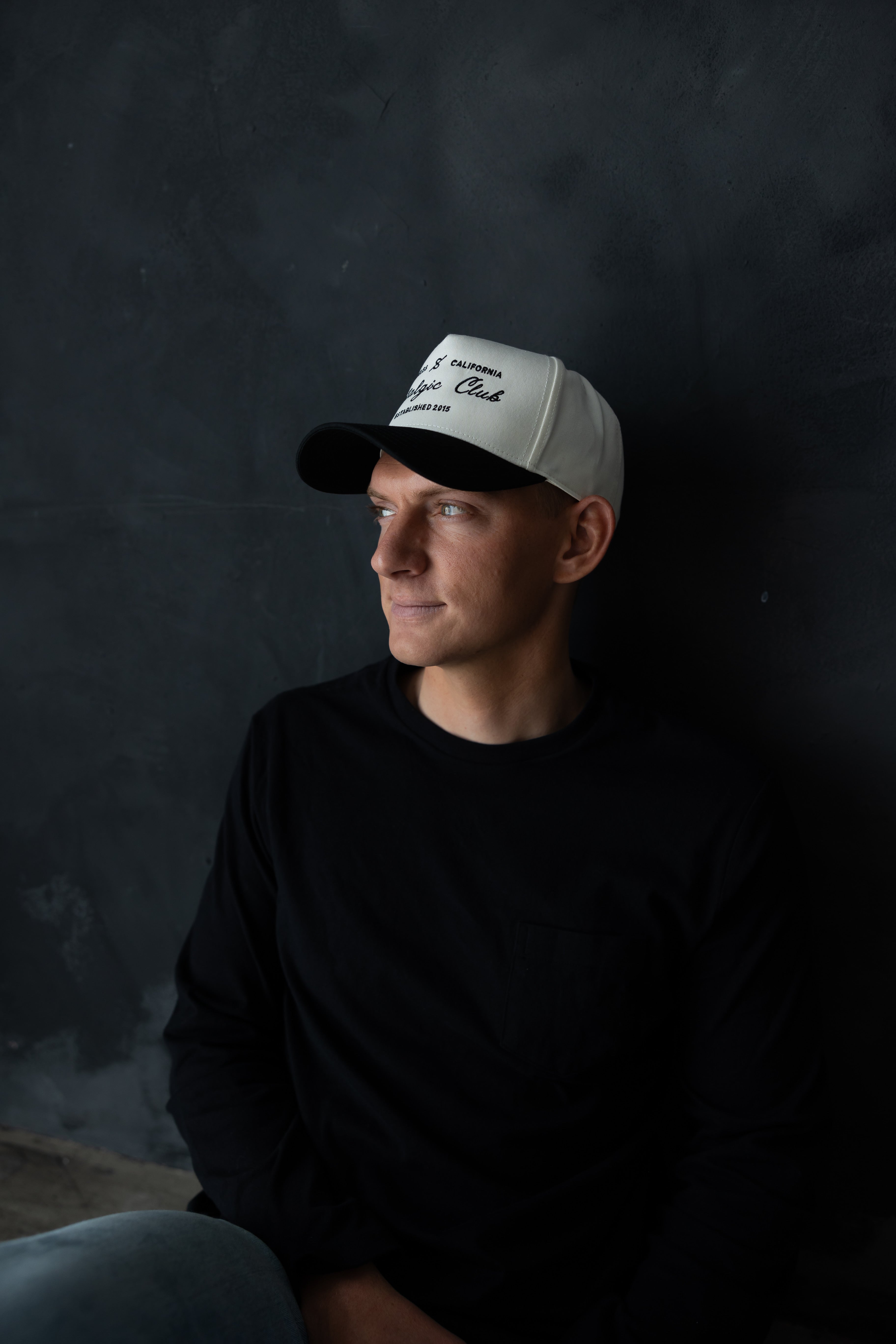 Man wearing Nostalgic Club black and white trucker hat by stitched apparel branded as stchd, styled with a minimalist black long-sleeve shirt, modern everyday look.