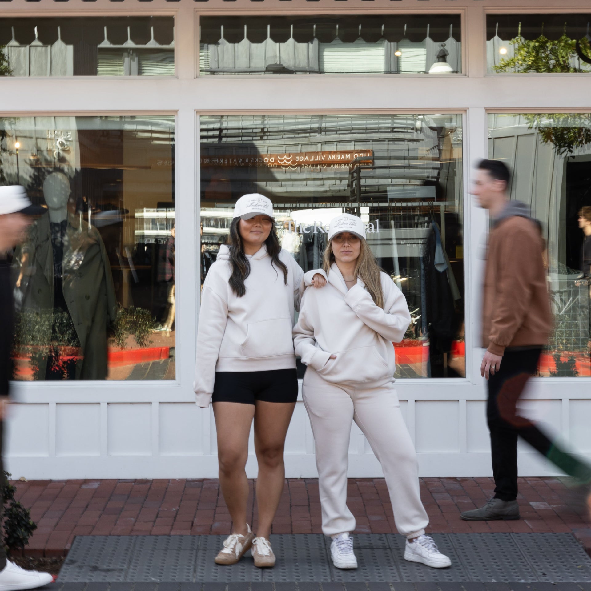 women wearing matching off-white oversized hoodies, one styled with sweatpants and the other with shorts, both accessorized with white embroidered caps. Versatile everyday essentials by Stitched Apparel branded as STCHD