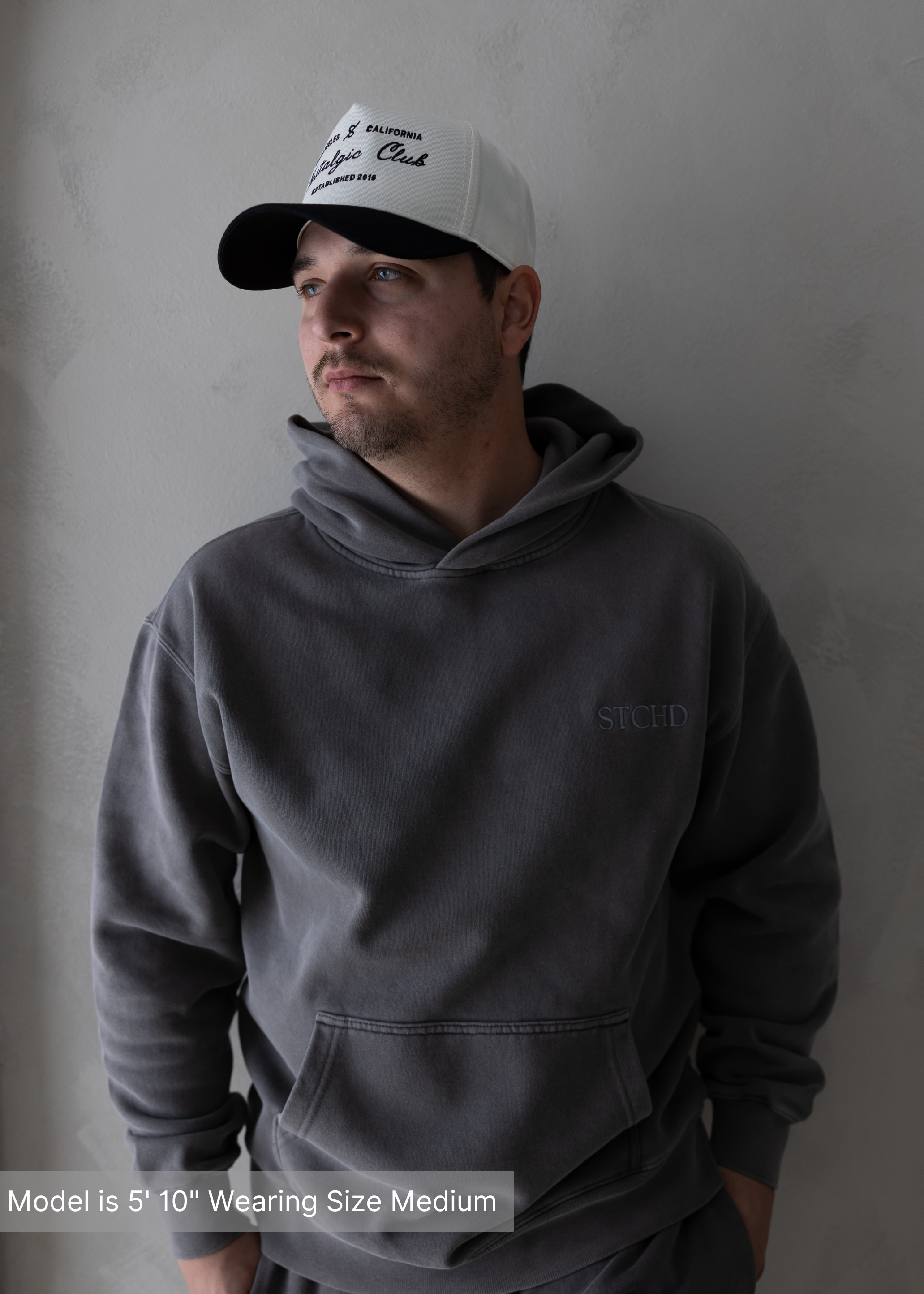 Man wearing washed charcoal hoodie and white Collective Club trucker hat, casual modern outfit.