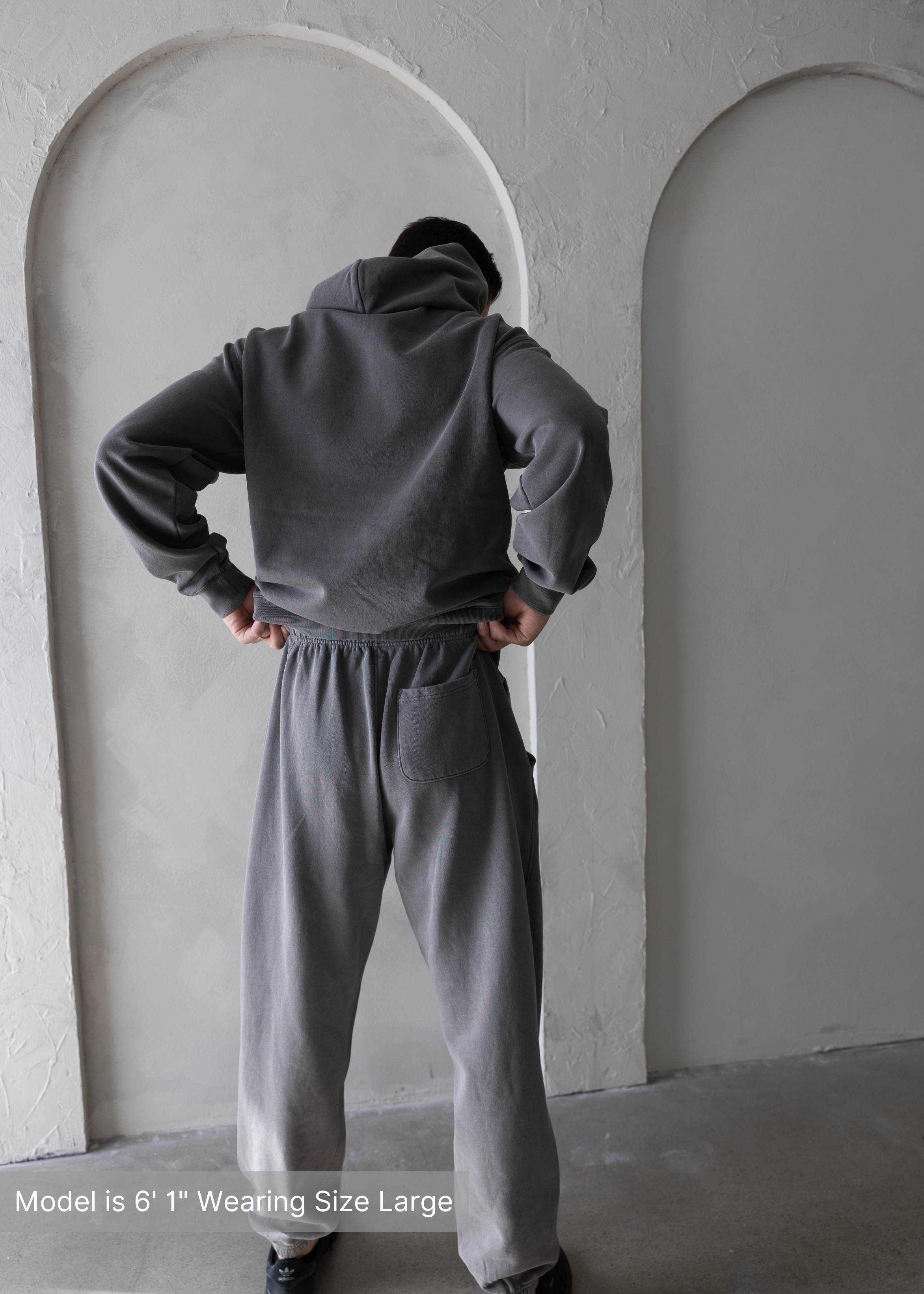 Back view of washed grey men’s hoodie and joggers set by stitched apparel branded as stchd, with hands on hips, relaxed athleisure outfit.