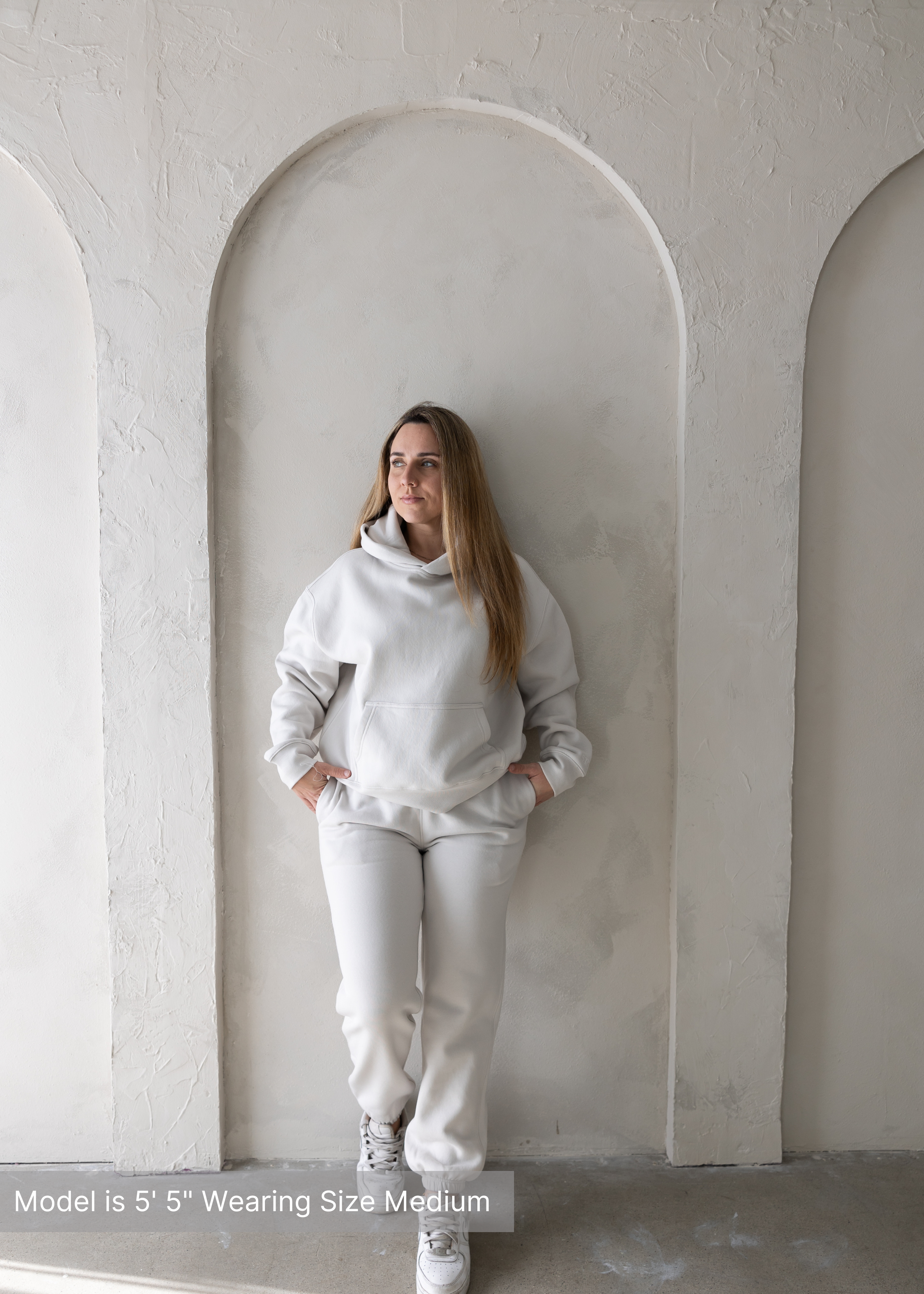 Woman wearing beige loungewear hoodie and sweatpants set by stitched apparel branded as stchd, front view posed against arched textured wall.