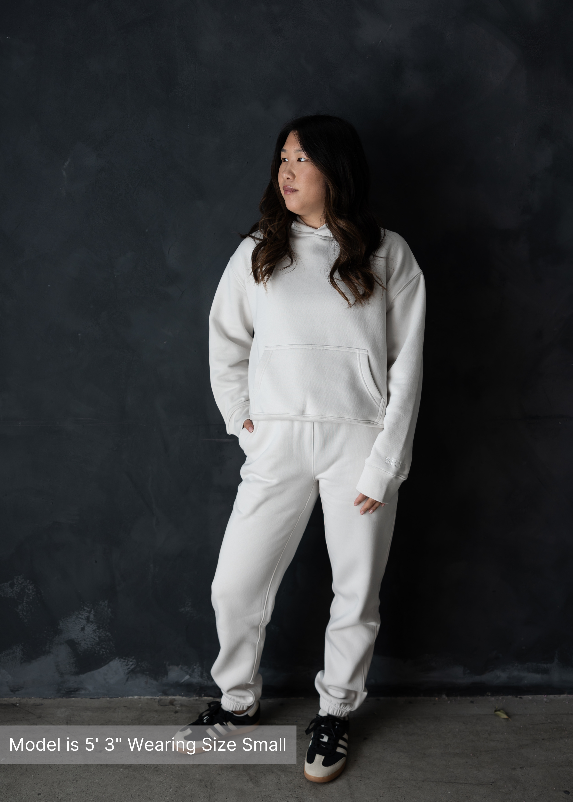 Woman wearing neutral loungewear set with sweatshirt and sweatpants by stitched apparel branded as STCHD, front view against black wall, model is 5'3" wearing size small.