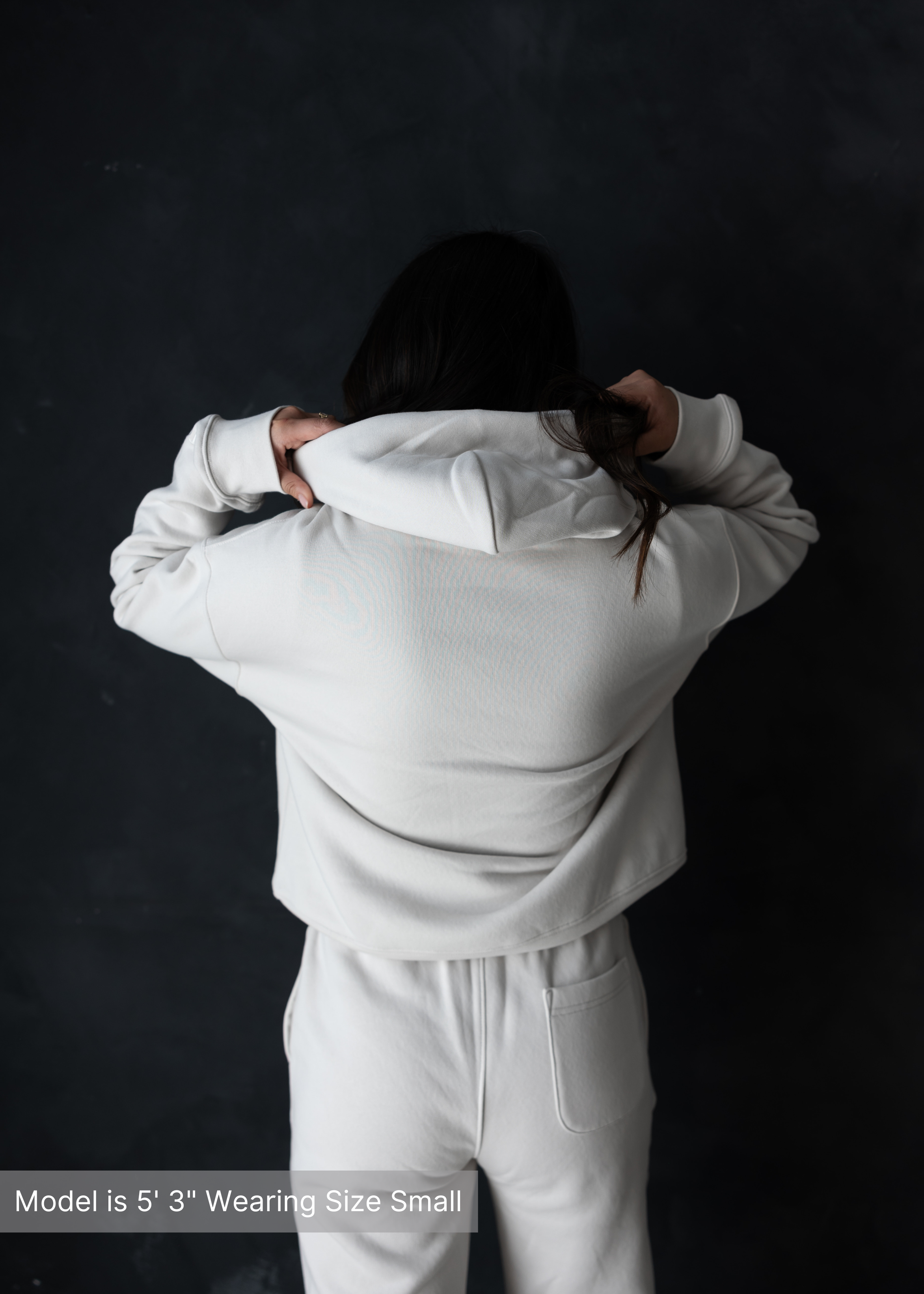 Woman lifting hoodie over her head, wearing neutral sweatsuit from behind, posed in front of dark wall.