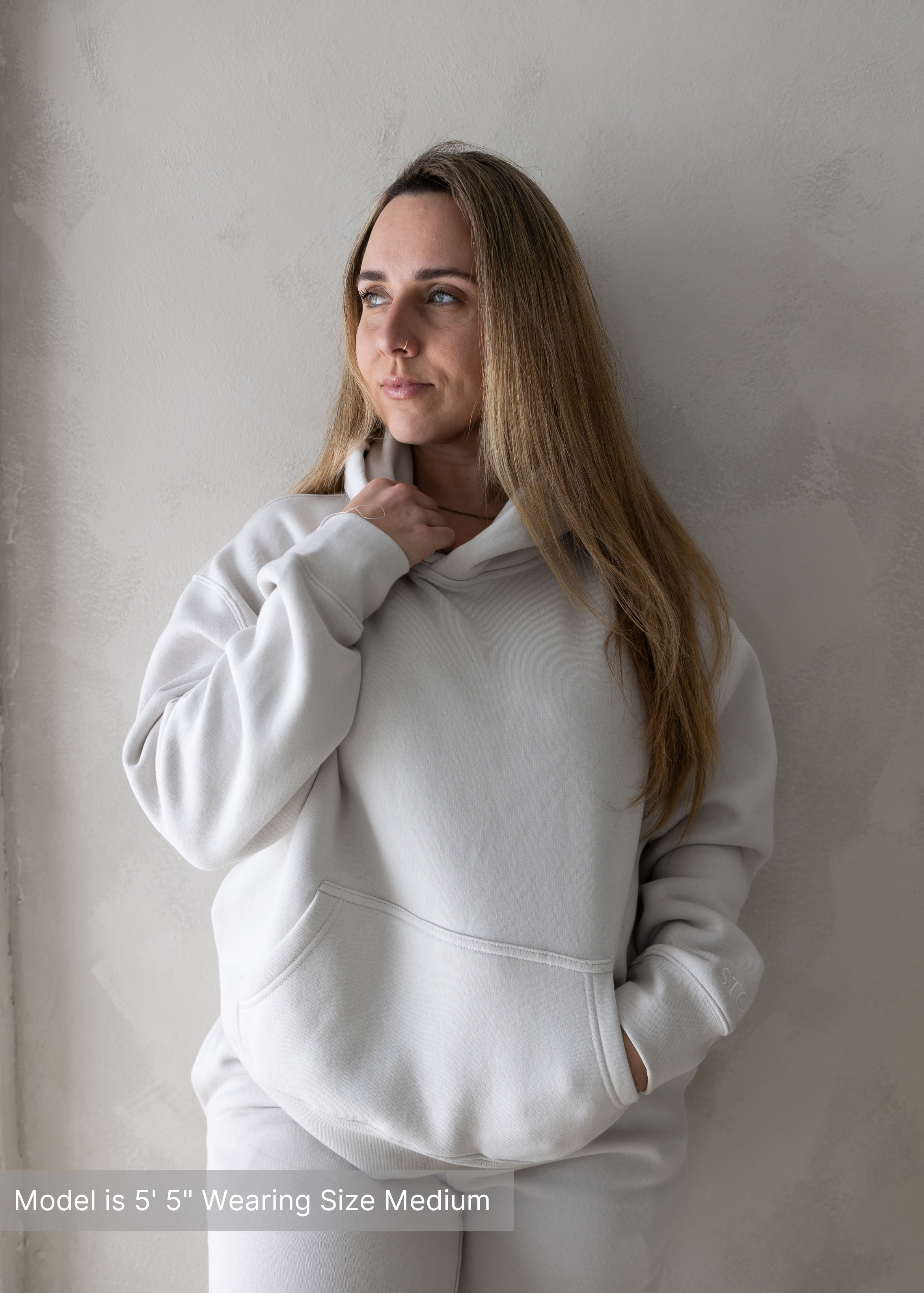 Woman wearing neutral hoodie and sweatpants set by stitched apparel branded as stchd, front profile with hand at collar, model is 5’5” wearing size medium